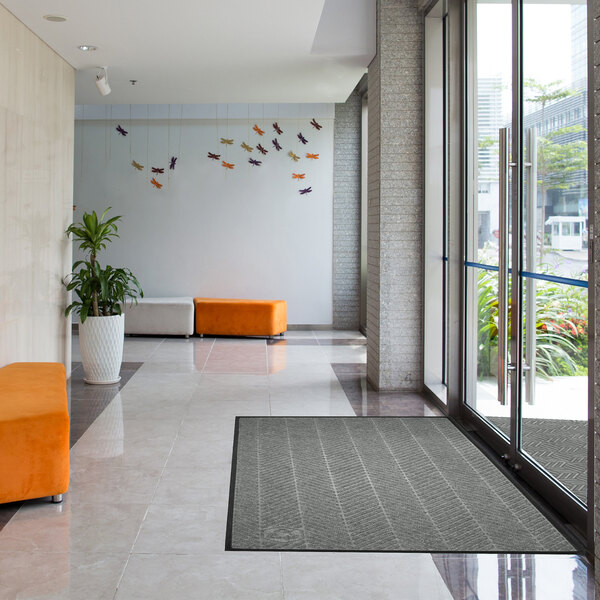 A modern lobby area with a large gray rectangular mat placed near glass entrance doors, featuring tiled floors, orange and white seating, a potted plant, and decorative wall art.