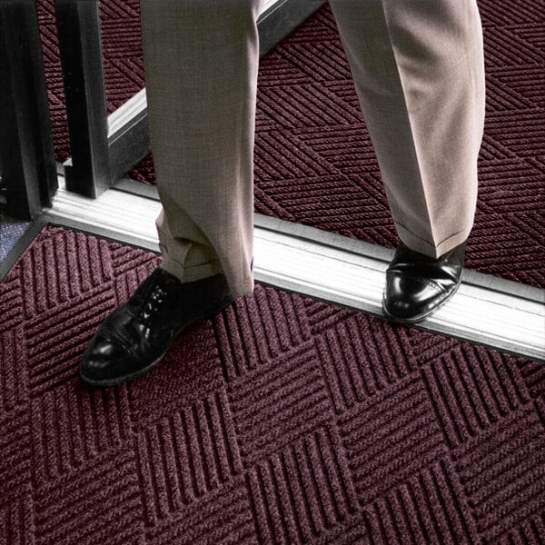A person wearing dress pants and black shoes is standing on a maroon textured mat with a geometric pattern near a doorway.