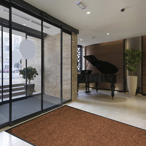 A large brown textured mat with a black border is placed on a light-colored floor near a glass entrance in a modern lobby with a grand piano and potted plants.