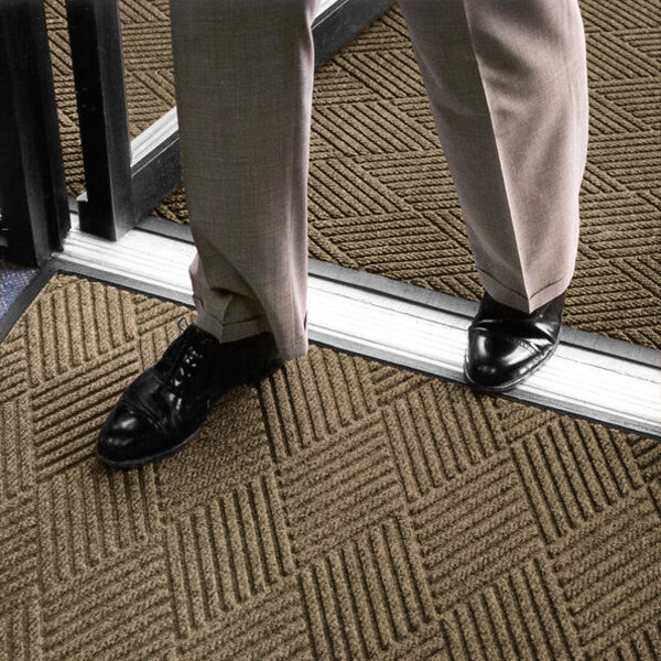 A person wearing light-colored pants and black shoes is standing on a textured, patterned brown mat near a doorway.