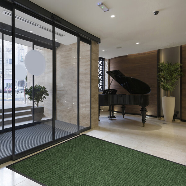 A modern lobby area with a large green mat on the floor, a grand piano in the background, and glass doors leading outside.