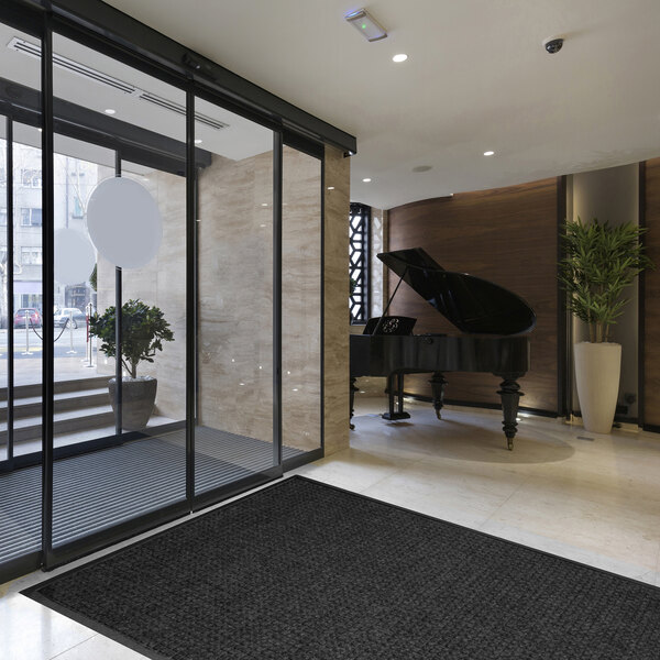 A modern lobby area with a large dark mat on the floor, a grand piano in the background, and glass doors leading outside.