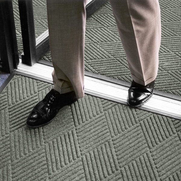 A person wearing dress pants and black shoes is standing on a textured gray mat with a geometric pattern near a doorway.