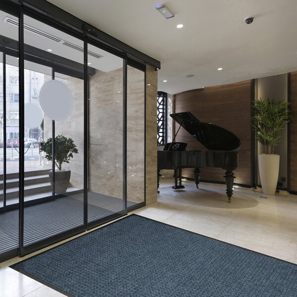 A modern lobby area with a large blue mat on the floor, a grand piano in the background, and glass doors leading outside.