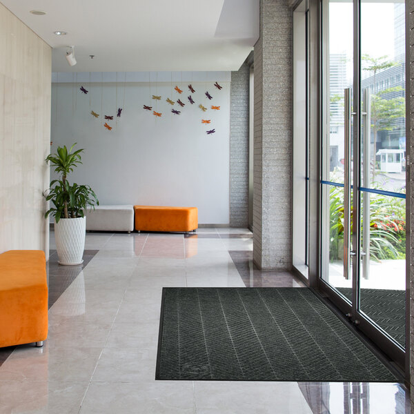 A black rectangular floor mat with a textured pattern is placed near the glass entrance doors of a modern lobby with tiled floors, orange and white seating, a potted plant, and decorative wall art.