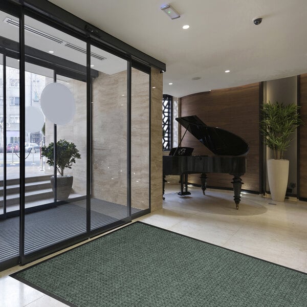 A modern lobby area with a large greenish mat on the floor, a grand piano, potted plants, and glass entrance doors.