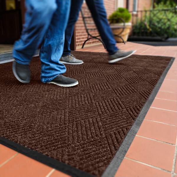 Two people are walking on a large, dark brown textured mat with a diamond pattern, placed on a tiled outdoor surface near a building entrance.