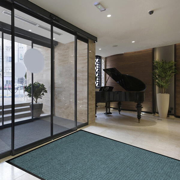 A modern lobby area with a large blue floor mat near glass doors, a grand piano, and potted plants.