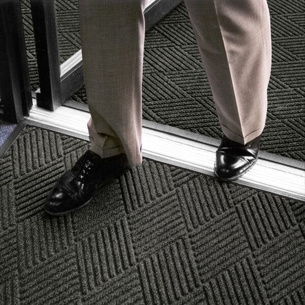 A person wearing dress pants and black shoes is standing on a textured, dark-colored mat with a geometric pattern near a doorway.