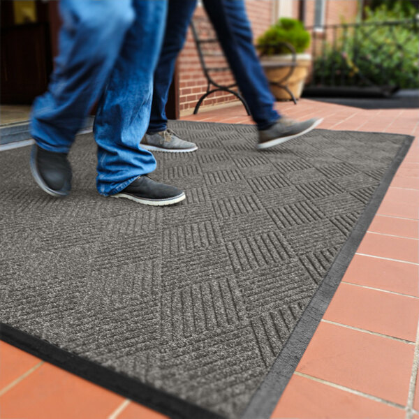 Two people are walking on a medium gray mat with a diamond pattern, placed on a tiled outdoor surface near a building entrance.