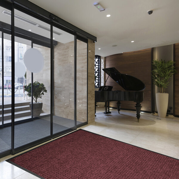 A modern lobby area with a large glass entrance, a red textured mat on the floor, a grand piano, and a potted plant.