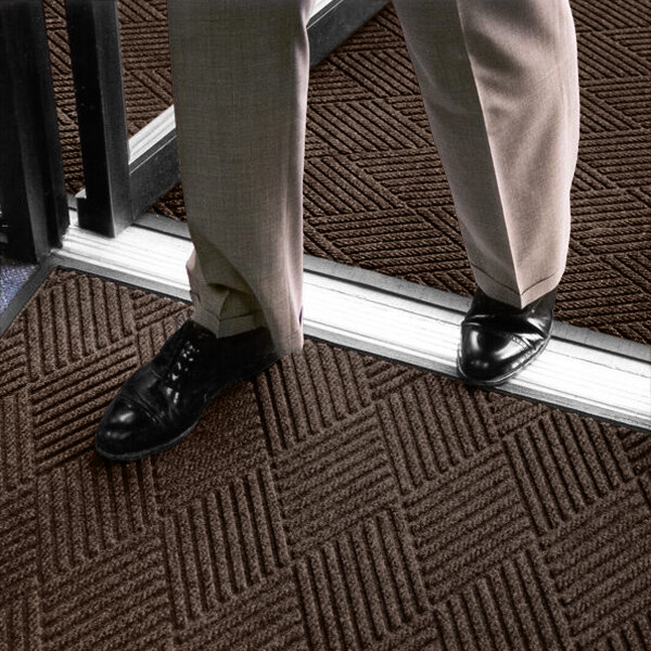 A person wearing dress pants and black shoes is standing on a textured brown mat with a geometric pattern near a doorway.