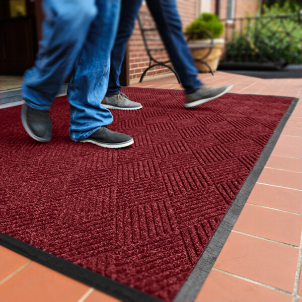 Two people are walking on a large red textured mat with a black border placed on a tiled outdoor surface.