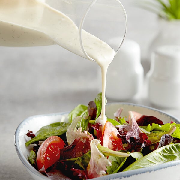 A bowl of mixed green salad with tomatoes being topped with creamy ranch dressing from a glass container.