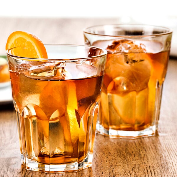 Two Fortessa Basics Chez Bistro rocks glasses of brown liquid with ice and orange slices on a table.