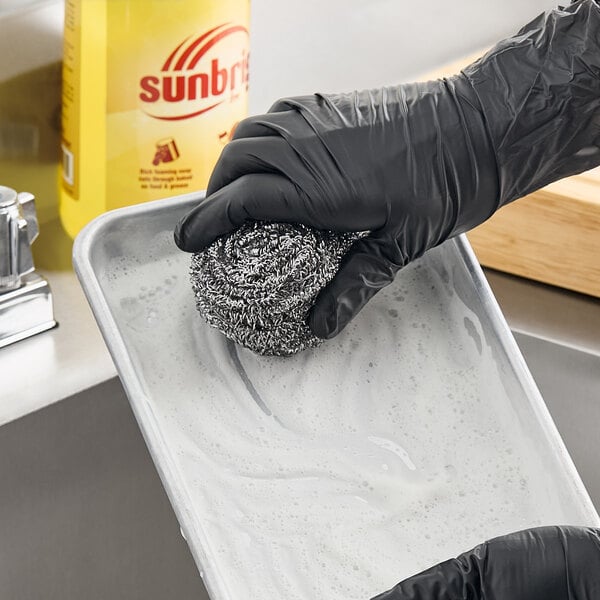 A stainless steel scrubber being used to clean a soapy metal tray.
