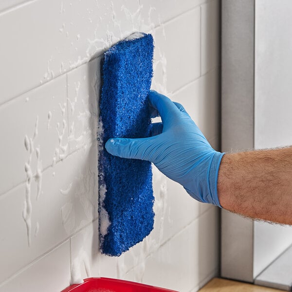 A hand in a blue glove using a Lavex Blue Multi-Purpose Scouring Pad to clean a wall.