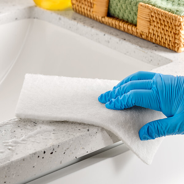 A hand in a blue glove using a Lavex white scouring pad to clean a counter.