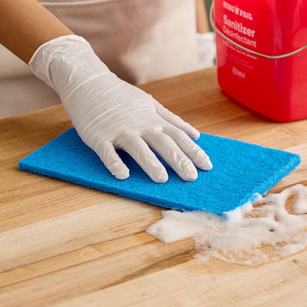 A person wearing white gloves cleaning a wooden table with a blue Lavex light-duty scouring pad.