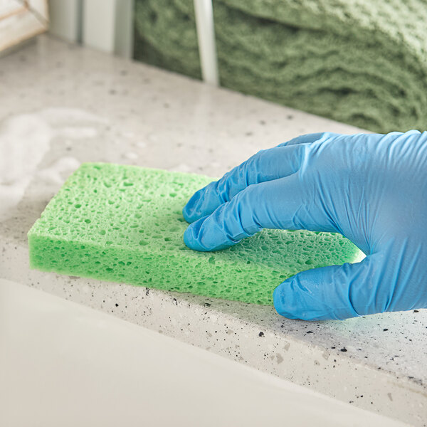A hand in a blue glove holding a green Lavex cellulose sponge cleaning a counter.