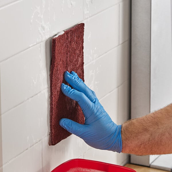 A hand wearing a blue glove uses a Lavex maroon scouring pad to clean a wall.