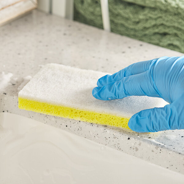 A gloved hand using a Lavex yellow and white sponge to clean a counter.