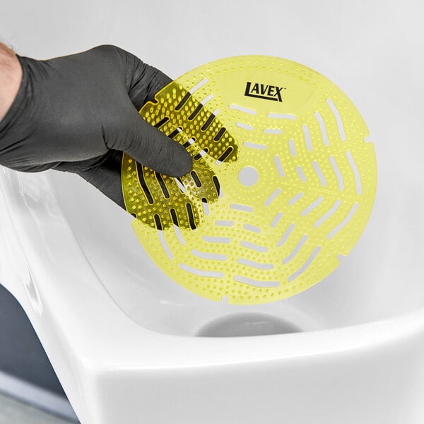 A hand in a black glove holding a yellow Lavex Mango Scent Deodorized Gel Urinal Screen.