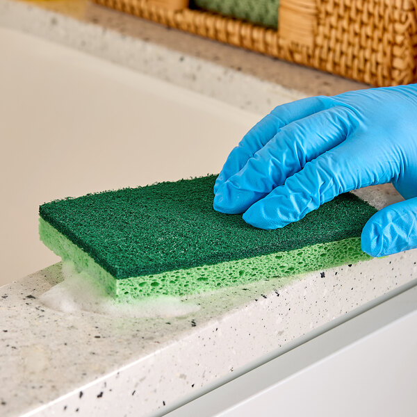 A hand in a blue glove using a green Lavex sponge to clean a counter.