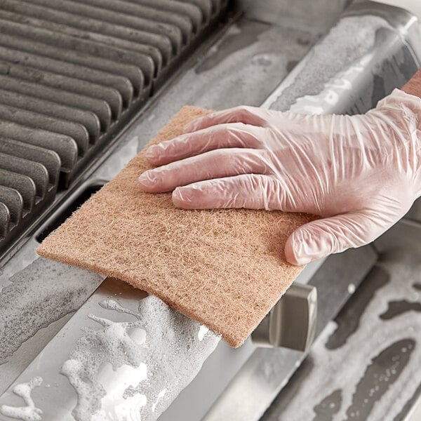 A gloved hand using a Lavex Eco Heavy-Duty Sisal Scouring Pad to clean a stove.