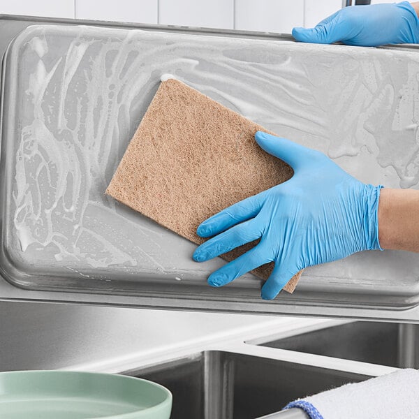 A gloved hand cleaning a tray with a Lavex Eco Heavy-Duty Sisal Scouring Pad.