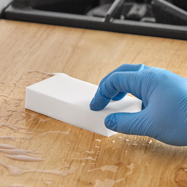A person in blue gloves using a white Lavex eraser sponge to clean a wooden table.