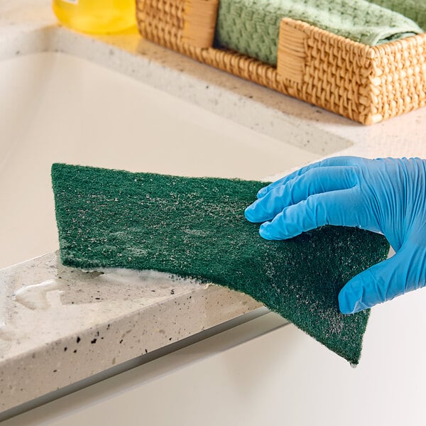 A gloved hand uses a Lavex dark green scouring pad to clean a countertop.