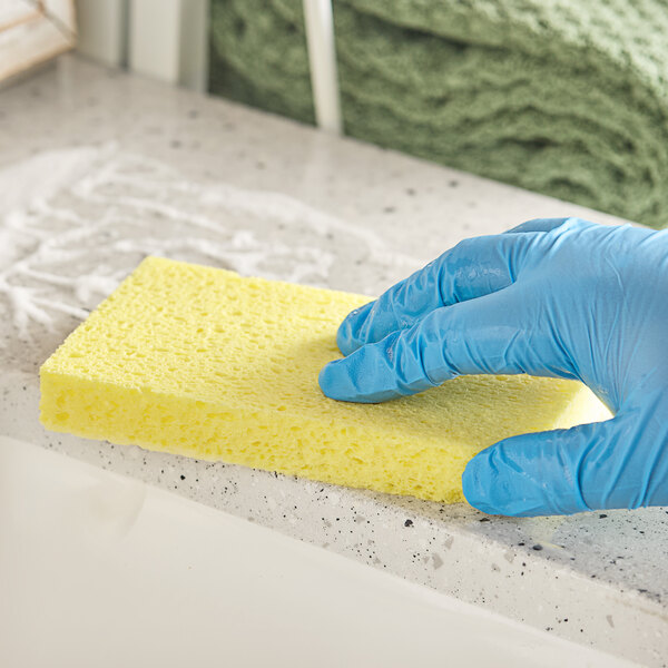 A hand in a blue glove cleaning a yellow sponge on a counter.