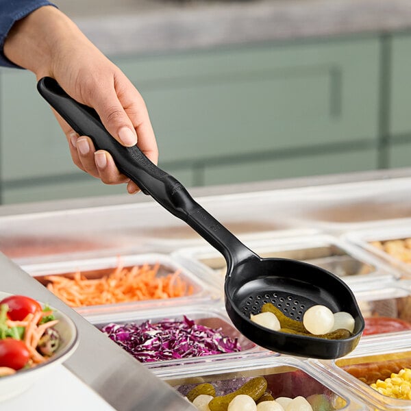 A hand holding a black Vollrath spoodle over a tray of vegetables.