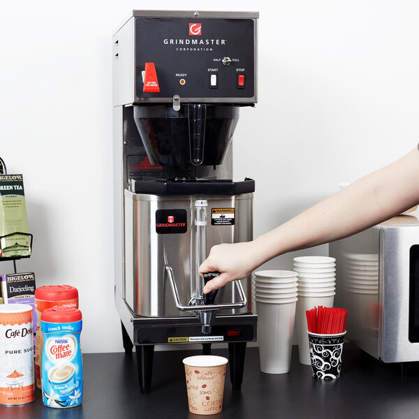 A person using a Grindmaster coffee machine to brew coffee into a white container.