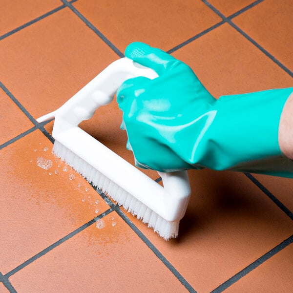 A hand in a green glove using a Carlisle white grout brush to clean tile.