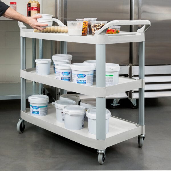 A person using a Rubbermaid three shelf utility cart to hold plastic containers of food.