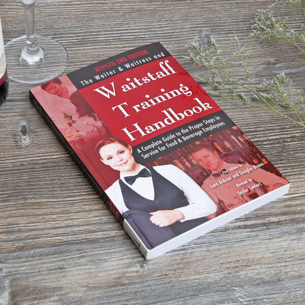 The Waiter, Waitress & Waitstaff Training Handbook on a table in a farm-to-table restaurant next to a glass of wine.