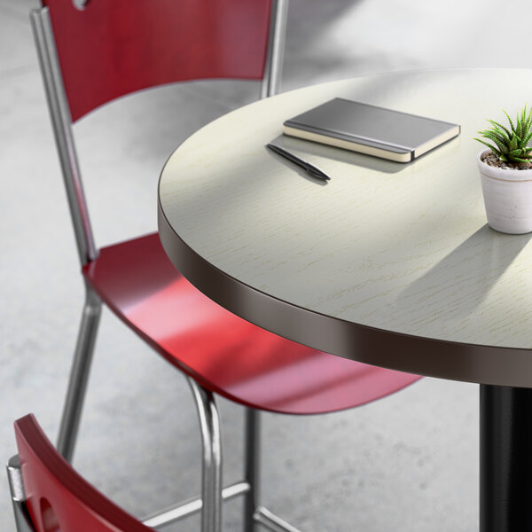 A round bar height table with a light wood laminate top, accompanied by red chairs, a small potted plant, a notebook, and a pen.