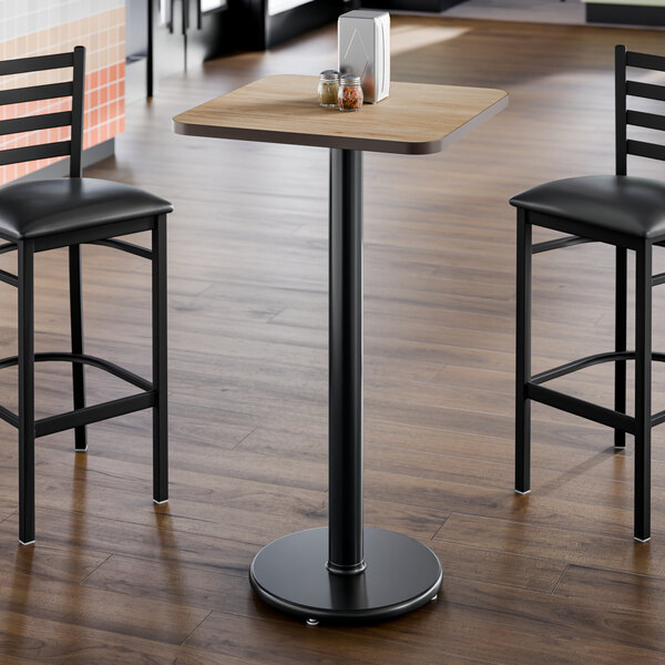 A square bar height table with a light wood laminate top and a black cast iron base.