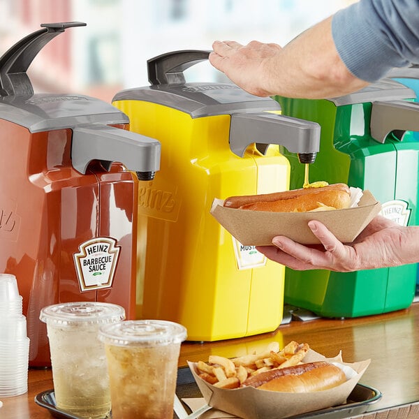 A person using a Heinz yellow plastic countertop mustard pump to pour mustard on a hot dog.