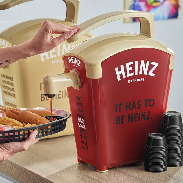 A person using a Heinz countertop pump to add ketchup to a basket of hot dogs.