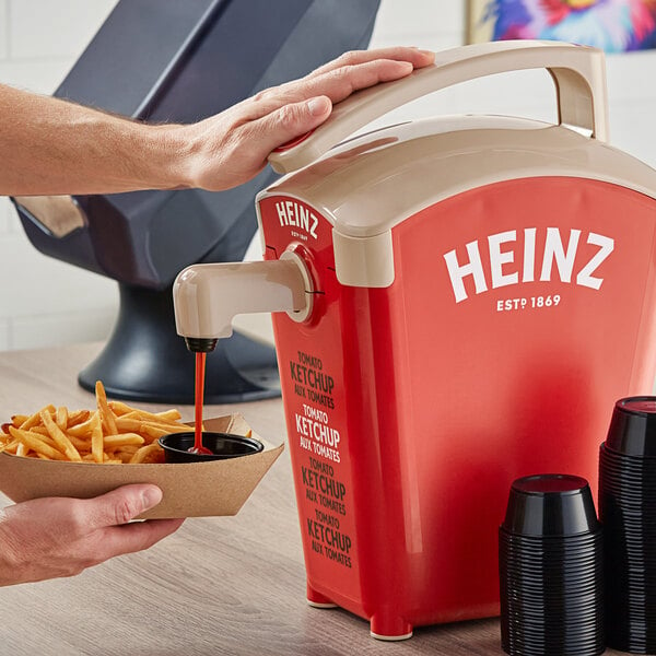 A person using a Heinz ketchup pump to pour ketchup into a black plastic cup of fries.