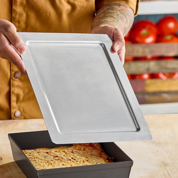 A rectangular metal deep dish pizza pan separator or lid being held above a pizza pan.