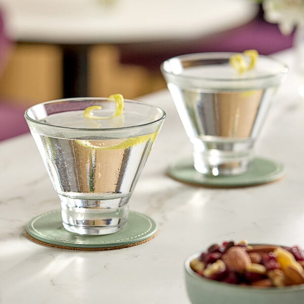 Two Libbey stemless martini glasses filled with a yellow liquid and garnished with lemon peel on a table with a bowl of nuts.