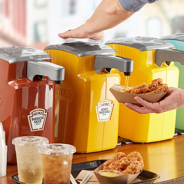 A person using a Heinz countertop pump to dispense mustard onto a plate of fried chicken.