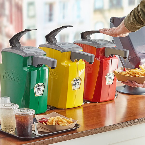 A person using Heinz red, yellow, and green countertop pump dispensers to add ketchup to a cup of fries.
