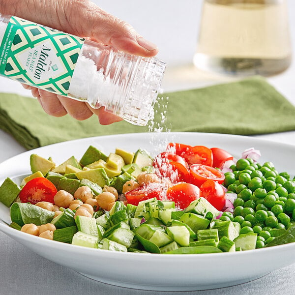 A hand using a Maldon Sea Salt grinder to sprinkle salt on a bowl of salad.