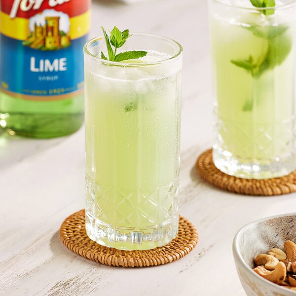 A glass of green limeade with mint leaves and a bowl of cashews on a table with a Torani Lime Flavoring bottle.