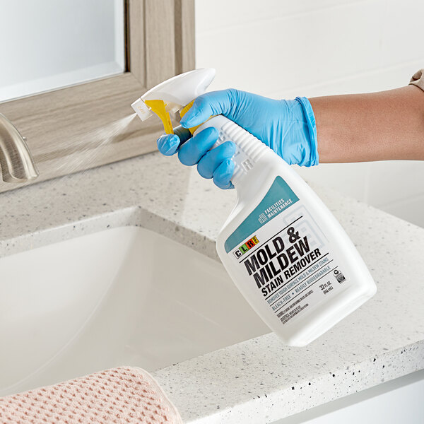 A person in blue gloves spraying CLR Mold & Mildew Stain Remover on a bathroom sink.